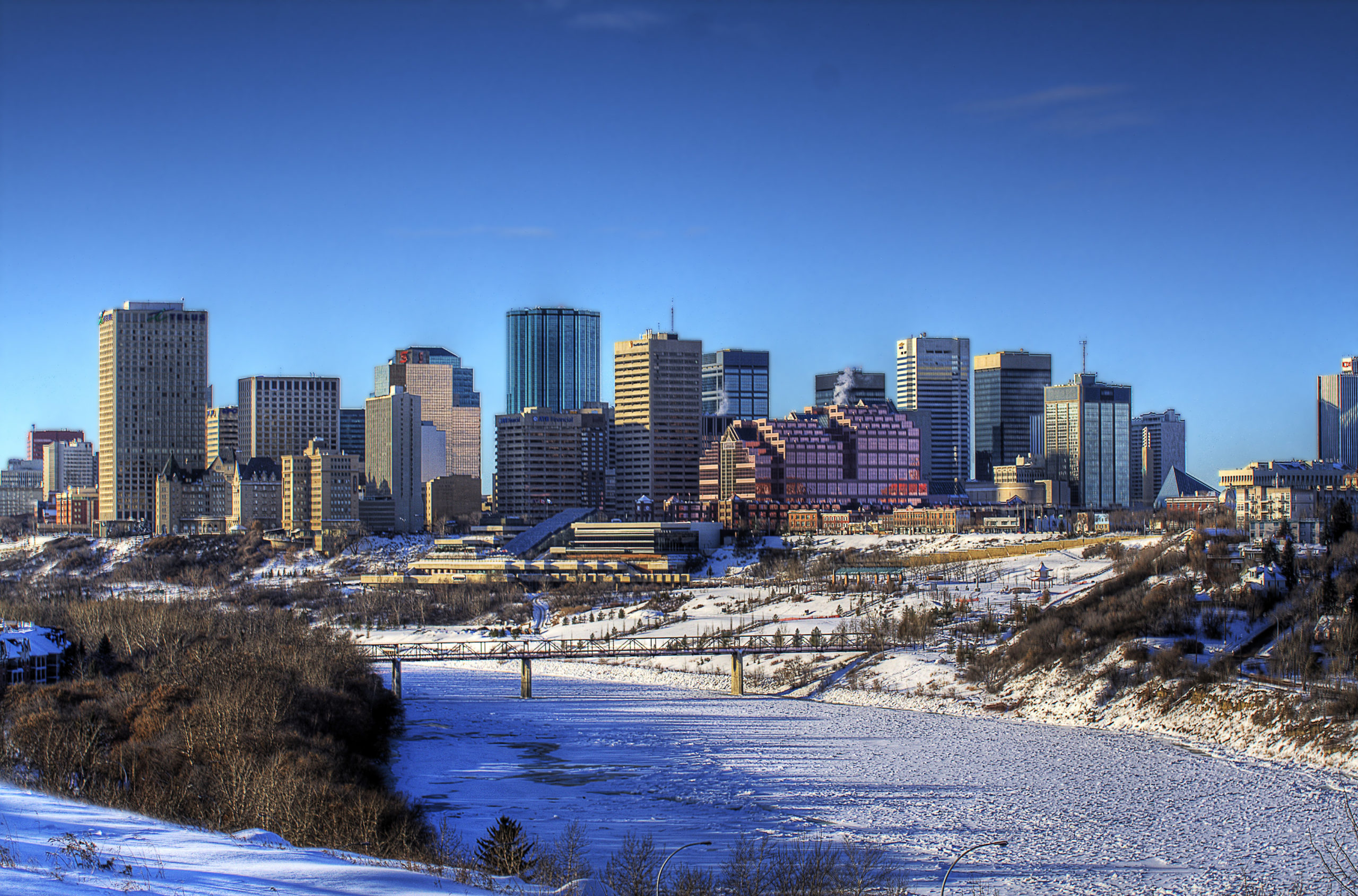 A photograph of the Edmonton skyline in winter with a blue sky and the frozen river in the foreground.